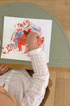 Photo prise en hauteur ou l'on appercçoit la main d'un enfant qui est entrain de faire de la peinture sur une feuille posé sur un set de table de couleur vert olive qui protège la table