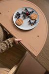 Photo d'un set de table en silicone marron posé sur une table en bois avec dessus une assiette contenant des biscuits et des myrtilles. On aperçoit également la main d'un enfant