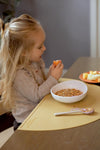 Jeune fille aux cheveux blonds et longs assise à table en train de manger son petit dejeuner, elle tient dans sa main un morceaux de fruit