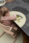 Jeune enfant assit à table grace à une chaise haute en bois, il a devant lui une assiette posé sur un set de table. Dans l'assiette, il y a des pâtes et de la salade. L'enfant est de dos et on le voit picorer dans l'assiette